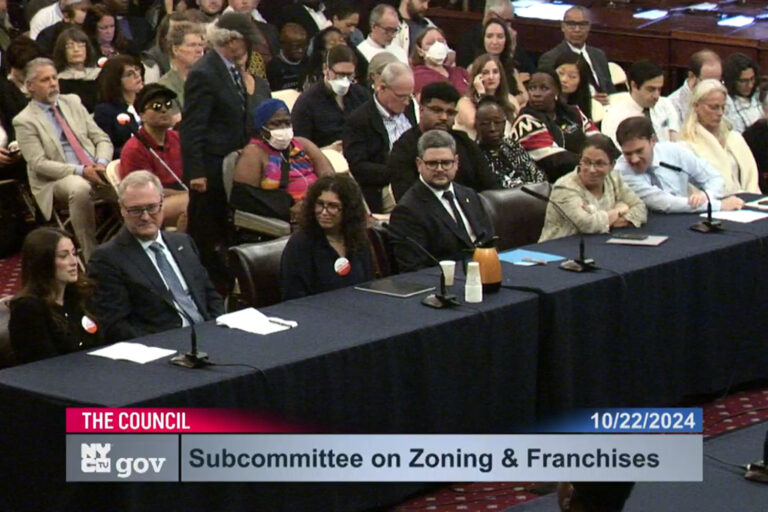 The Yes to Housing hearing. Policy makers sit at a black table, with citizens sitting in chairs behind them.