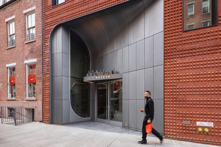 Exterior shot of the Italian Art Museum entrance point, a gray perforated metal facade. A person walks along the sidewalk.