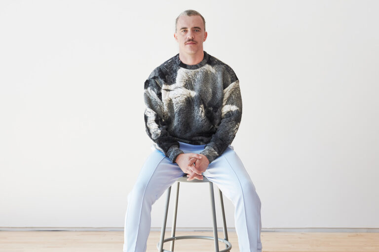 Portrait of Peter Zuspan, a white man with short dark hair wearing a gray and white graphic sweater and powder blue pants. He sits on a stool against a white background.