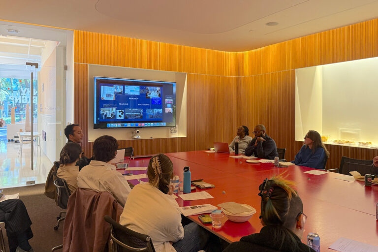 A group sits around a large red table looking at a screen on the wall showing a Zoom meeting.