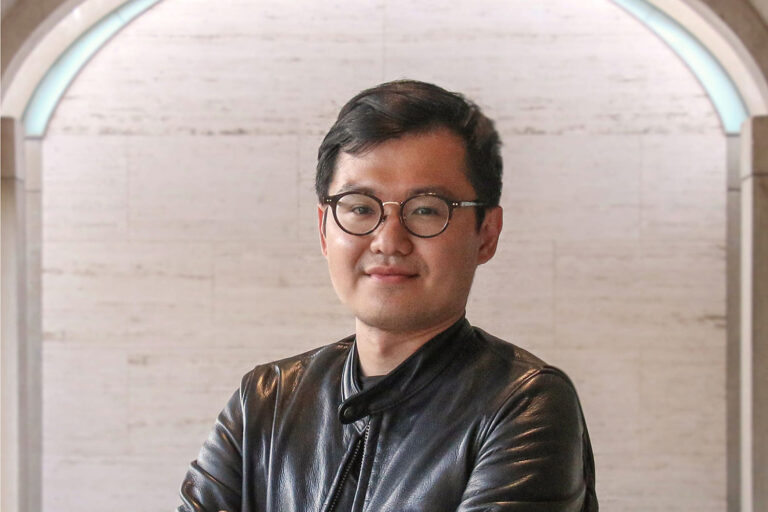 Portrait of an adult Asian man with short black hair, wearing black, round-framed glasses.