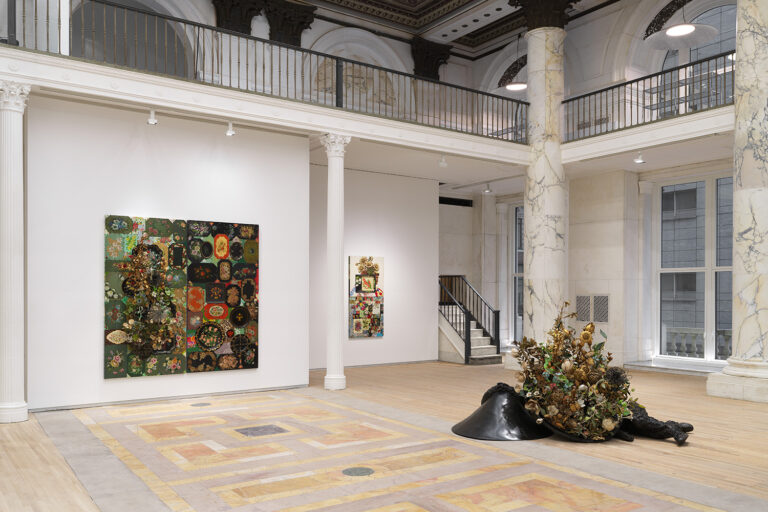 Interior scene of art gallery with paintings on white walls and a sculpture on the ground.